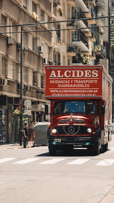 A red moving truck with the company name 'Alcides' and advertised services for furniture transport and storage is parked on a city street in Maida Vale, London. The truck is positioned facing forward, occupying a lane next to a pedestrian crossing. Behind the truck, multi-storey residential buildings with balconies and small potted plants are visible, with some windows open and air conditioning units attached. To the left of the truck, a person wearing dark clothing and a face mask stands on the pavement, near a bicycle. The street is lined with shops and apartment entrances, with a few more pedestrians walking nearby. The lighting is bright, indicating daytime, and the scene captures a typical urban environment suitable for home relocation and furniture transport logistics carried out by Maida Vale Man and Van, demonstrating the loading process of household goods onto the vehicle.