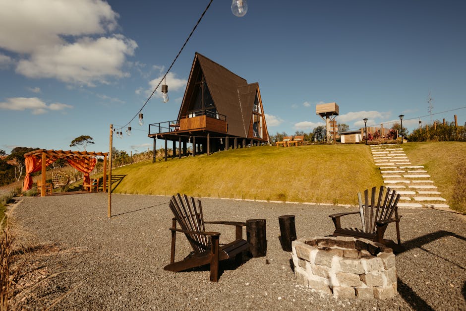The image shows an outdoor scene with a modern A-frame wooden house elevated on stilts, located on a grassy hill under a partly cloudy sky. In the foreground, there is a gravel area featuring two wooden Adirondack chairs positioned around a small circular stone fire pit. To the left, a wooden pergola with orange fabric drapes provides a shaded seating area, and nearby, a tall wooden pole supports a string of outdoor hanging lights. On the right side, a concrete staircase with white-painted edges leads up the grassy slope toward the house. The setting includes various outdoor furniture pieces, such as benches and a picnic table, suggesting an area designed for relaxation and outdoor leisure, typical of a home relocation or property setup. The lighting indicates natural daylight, with shadows cast by the furniture and structures, emphasizing the scene's clarity. This outdoor arrangement could be part of a relocation setup managed by companies like Maida Vale Man and Van, supporting home moving or furniture transport activities.