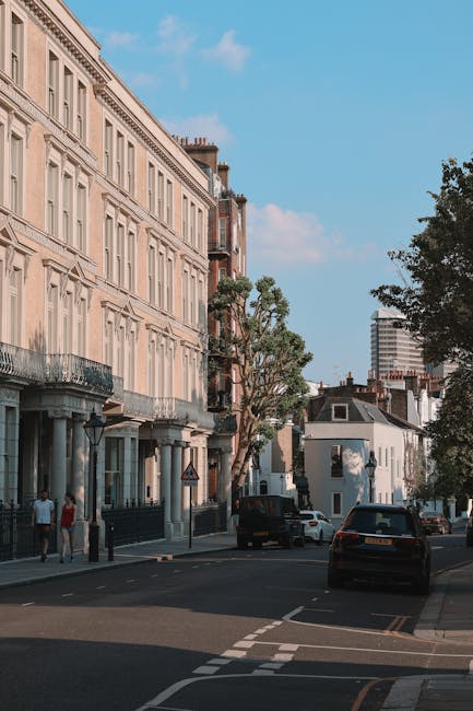 A red moving truck with the company name 'Alcides' and advertised services for furniture transport and storage is parked on a city street in Maida Vale, London. The truck is positioned facing forward, occupying a lane next to a pedestrian crossing. Behind the truck, multi-storey residential buildings with balconies and small potted plants are visible, with some windows open and air conditioning units attached. To the left of the truck, a person wearing dark clothing and a face mask stands on the pavement, near a bicycle. The street is lined with shops and apartment entrances, with a few more pedestrians walking nearby. The lighting is bright, indicating daytime, and the scene captures a typical urban environment suitable for home relocation and furniture transport logistics carried out by Maida Vale Man and Van, demonstrating the loading process of household goods onto the vehicle.