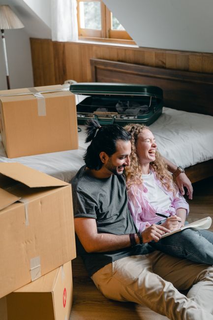 A young couple sitting on the floor of a room during a home relocation process, surrounded by open cardboard boxes, some sealed with packing tape, and a dark green suitcase with an open lid revealing 