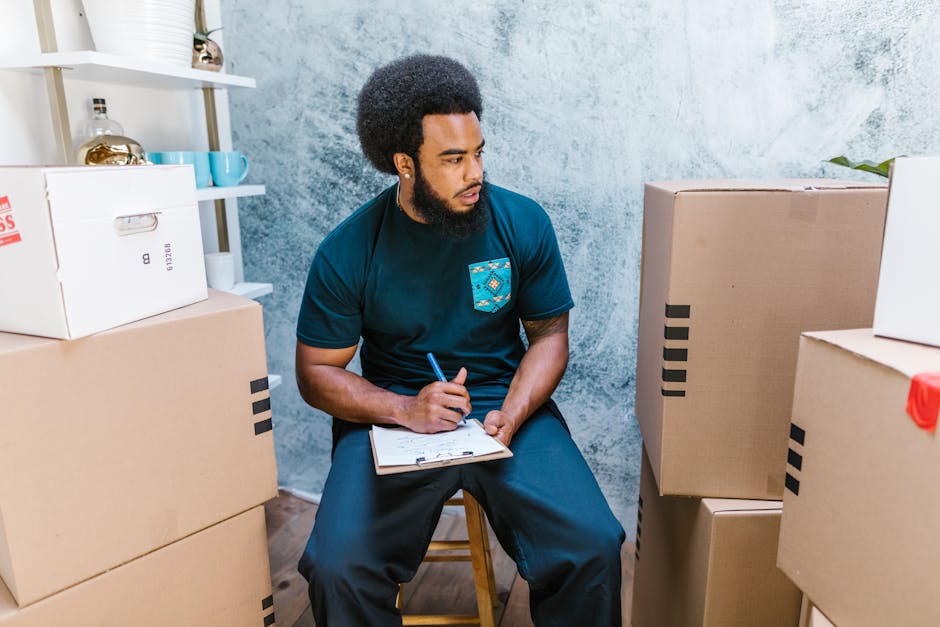 A man with an afro hairstyle and a beard is sitting on a wooden stool inside a room with textured light blue walls. He is wearing a dark blue t-shirt with a patterned pocket and holding a clipboard and a pen in his right hand, appearing to be planning or taking notes. Around him are several large cardboard boxes, some sealed with packing tape, placed on the floor and stacked against the wall, likely containing household items for a home relocation. The room contains white shelving with decorative objects, including a golden vase and a ceramic mug. Natural light illuminates the scene, emphasizing the packing and moving process. The image visually represents the preparation phase of furniture transport and packing during a house move, aligning with services provided by Maida Vale Man and Van in their removals category, with a focus on efficient and organized household logistics.