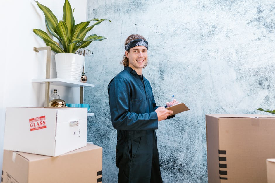 A man with an afro hairstyle and a beard is sitting on a wooden stool inside a room with textured light blue walls. He is wearing a dark blue t-shirt with a patterned pocket and holding a clipboard and a pen in his right hand, appearing to be planning or taking notes. Around him are several large cardboard boxes, some sealed with packing tape, placed on the floor and stacked against the wall, likely containing household items for a home relocation. The room contains white shelving with decorative objects, including a golden vase and a ceramic mug. Natural light illuminates the scene, emphasizing the packing and moving process. The image visually represents the preparation phase of furniture transport and packing during a house move, aligning with services provided by Maida Vale Man and Van in their removals category, with a focus on efficient and organized household logistics.