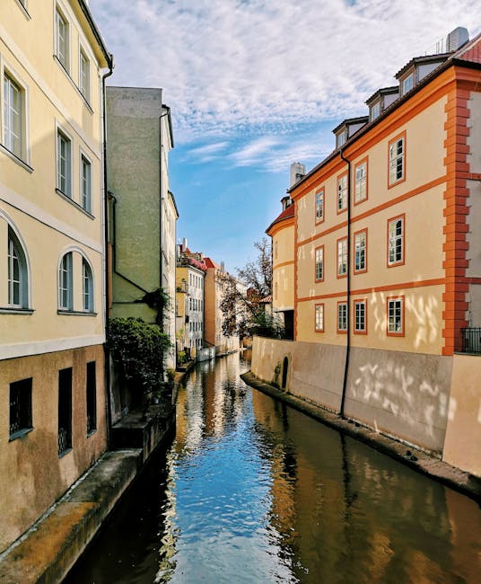 A narrow canal lined with multi-storey residential buildings on both sides is visible in the image. On the left, a pale yellow building features multiple windows with white frames and small arched window openings near the ground level. On the right, an orange-brick apartment building with white window frames and a black drainpipe runs down its facade. The canal water reflects the surrounding buildings and the partly cloudy sky overhead. The scene is illuminated by natural daylight, highlighting the textures of the brickwork and plastered walls. The canal walls are made of concrete with some moss and dirt at the waterline. Tree branches extend into the frame from the left side, and a small bridge or pathway might be just out of view. This urban setting depicts a typical scene of a home relocation area, with the buildings and canal emphasizing the logistical considerations involved in furniture transport and packing during a house move. The image is typical of the Little Venice area of Maida Vale, where Maida Vale Man and Van offers professional removals and moving services.