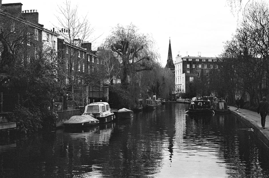 Black and white photograph of a narrow waterway in Maida Vale, with several moored boats along the banks, some covered with tarps. On the left side, a row of multi-story residential buildings with brick facades and chimneys extend into the distance. Bare trees line the canal, with one large tree in the center having prominent branches and no leaves. In the background, there is a tall church spire rising above the buildings. On the right side, two people are walking along the pavement, and a van is parked partly visible behind the boats. The overall scene depicts an urban residential area with calm water reflecting the surrounding structures and trees, fitting the context of house removals and container transport in a peaceful canal setting. This image is used on MaidValemanandvan.co.uk's Little Venice Maida Vale removals and packing guide page to illustrate moving logistics in this historic canal location, emphasizing careful loading and transportation of household belongings by professional movers such as Maida Vale Man and Van.