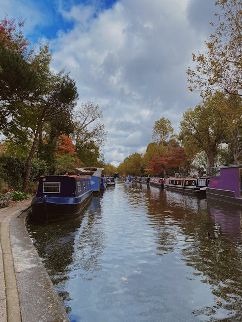 A narrow canal lined with multi-storey residential buildings on both sides is visible in the image. On the left, a pale yellow building features multiple windows with white frames and small arched window openings near the ground level. On the right, an orange-brick apartment building with white window frames and a black drainpipe runs down its facade. The canal water reflects the surrounding buildings and the partly cloudy sky overhead. The scene is illuminated by natural daylight, highlighting the textures of the brickwork and plastered walls. The canal walls are made of concrete with some moss and dirt at the waterline. Tree branches extend into the frame from the left side, and a small bridge or pathway might be just out of view. This urban setting depicts a typical scene of a home relocation area, with the buildings and canal emphasizing the logistical considerations involved in furniture transport and packing during a house move. The image is typical of the Little Venice area of Maida Vale, where Maida Vale Man and Van offers professional removals and moving services.