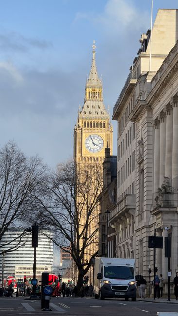 The image shows a city street scene in Maida Vale with a view of the Elizabeth Tower, commonly known as Big Ben, in the background under a partly cloudy sky. In the foreground, there is a white van parked on the pavement near a building with classical architectural features, including columns and decorative stonework. Trees without leaves are positioned along the street, indicating it might be late autumn or winter. The scene includes traffic lights, road signs, and a few pedestrians walking on the sidewalk. The Van is part of the loading process for a home relocation or furniture transport, relevant to the legal parking restrictions in Maida Vale, with the scene captured during daylight. Maida Vale Man and Van can be involved in such logistics, providing removals or packing and moving services in the area. The overall environment is urban, with a balance of historic and modern elements visible.