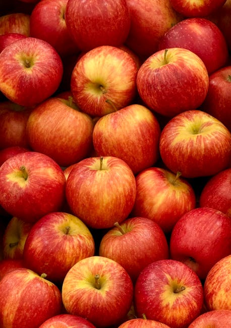 A close-up view of numerous red and yellow apples with smooth, shiny skins, piled together in a market or storage setting. The apples vary in size and have natural color variations, with some showing faint streaks of green or orange. They are arranged closely, filling the frame almost entirely, with no visible packaging or containers. The lighting highlights their glossy surface, emphasizing their freshness. This image captures the visual appeal of fresh apples, which are often involved in food-related activities or grocery shopping, related to house removals or packing and moving services that include handling and transporting fresh produce.