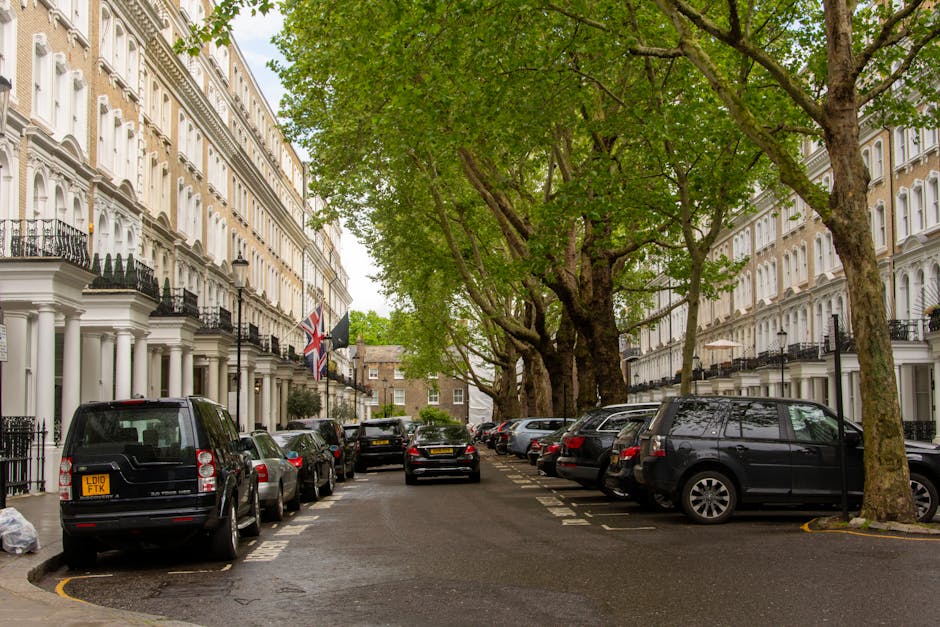 The image shows a city street scene in Maida Vale with a view of the Elizabeth Tower, commonly known as Big Ben, in the background under a partly cloudy sky. In the foreground, there is a white van parked on the pavement near a building with classical architectural features, including columns and decorative stonework. Trees without leaves are positioned along the street, indicating it might be late autumn or winter. The scene includes traffic lights, road signs, and a few pedestrians walking on the sidewalk. The Van is part of the loading process for a home relocation or furniture transport, relevant to the legal parking restrictions in Maida Vale, with the scene captured during daylight. Maida Vale Man and Van can be involved in such logistics, providing removals or packing and moving services in the area. The overall environment is urban, with a balance of historic and modern elements visible.