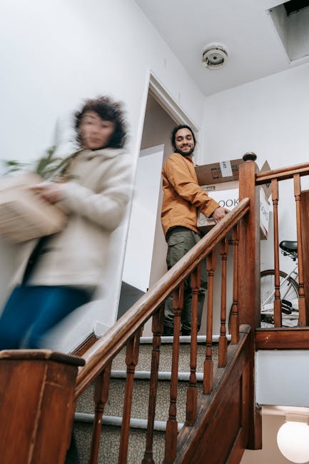 Inside a residential property, a man with a beard, wearing a brown jacket and dark trousers, stands on a staircase landing, smiling as he holds a cardboard box labeled 'BOOKS'. In the foreground, a woman dressed in a beige coat and blue jeans, with curly hair, is captured in motion as she walks past, carrying another cardboard box. The scene appears to be part of a home relocation or furniture transport process organized by Maida Vale Man and Van, with the staircase made of wood, featuring a polished banister, and the interior illuminated by natural light and ceiling fixtures. Visible packing materials include cardboard boxes, some marked for packing, and the overall environment suggests an active moving day with furniture and boxes staged for transport, potentially taking place during a loading process for a house removal service.