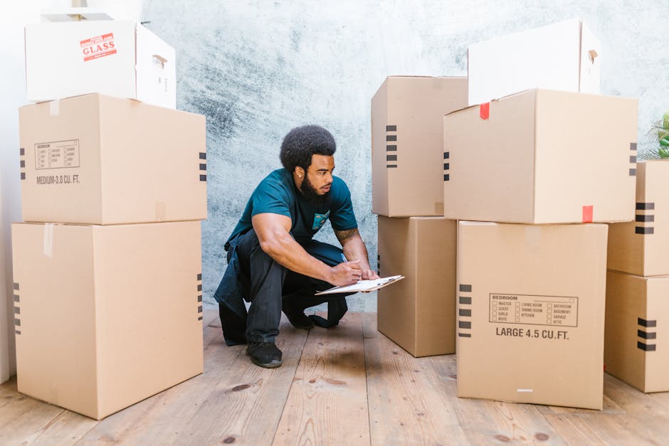 A man crouches on a wooden floor inside a property, surrounded by multiple large cardboard moving boxes with printed labels indicating contents such as 'bedroom' and 'large 4.5 cu ft.' He is holding a clipboard and appears to be inspecting or noting details during a home relocation process. The boxes are stacked against a plain, light-colored wall, with some positioned near a doorway or entrance. The scene is well-lit, emphasizing the packing and organization aspects associated with furniture transport and packing and moving activities. This image, representing the logistics involved in house removals, aligns with Maida Vale Man and Van's services, illustrating the careful handling, packing materials like cardboard and tape, and the loading process essential for professional removals.