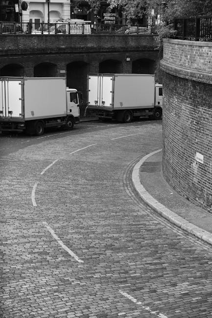 A black-and-white photograph captures a narrow, curved cobbled street in an urban residential area, with a brick wall on the right side and a metal railing above, overlooking a background of houses and trees. Two white moving trucks are parked along the street, with the rear sections facing the viewer, indicating the loading process. In the foreground, a small section of the cobbled pavement shows faint white dashed lines suggesting parking or loading zones. The scene illustrates a home relocation or furniture transport situation, typical of a house removals service like Maida Vale Man and Van, with the trucks positioned near an entrance for efficient loading and unloading of furniture and packed boxes. The environment appears calm, with soft lighting and no visible pedestrians or moving staff, focusing on the logistical aspect of moving services in the context of house removals.