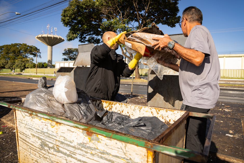 Two men are engaged in waste disposal outside a property during a house removal or packing process. One man, dressed in a grey T-shirt, is holding a cardboard box containing various items, while the other, wearing a black jacket and yellow gloves, is assisting by placing the box into a large, rusted metal dumpster. In the foreground, the open dumpster is filled with black and white plastic garbage bags, with some trash visible on top. The scene takes place on a paved street with a row of trees and a distinctive water tower in the background under a clear blue sky. Nearby, there is a curb and a sidewalk, and the property's doorway can be seen behind the men. The environment suggests a residential or suburban area, and the activity appears to be part of a home relocation or furniture transport process managed by Maida Vale Man and Van, aligning with local waste disposal regulations and moving logistics.