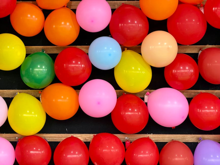 A collection of colourful balloons in shades of red, pink, yellow, green, orange, blue, and purple are arranged on a black surface with wooden slats, likely for a celebration or event. The balloons are inflated and glossy, reflecting light, and are positioned close together without overlapping. Some balloons appear to be resting on or near a delivery area, which could relate to the logistics of home relocation or packing and moving services. The setting suggests an indoor environment with bright, even lighting, and the arrangement may be part of pre-move decorations or packing materials for a residential move. Maida Vale Man and Van, a professional removals company, may use such vibrant displays when preparing for house removals or packing services in the Maida Vale area, emphasizing efficient and organized furniture transport and moving logistics.