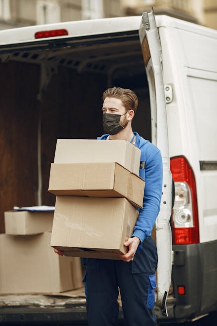 A man wearing a black face mask and a blue hoodie is standing outside a white commercial van with its rear door open. He is holding three cardboard boxes of varying sizes, stacked one on top of another, as part of a house removal or home relocation process. Inside the van, several additional cardboard boxes are visible, some sealed with packing tape and others open, revealing packing materials like paper or plastic wrapping. The scene depicts the loading process involved in furniture transport and packing during a house move in Maida Vale. The natural lighting highlights the details of the cardboard boxes, the man's attire, and the vehicle's interior, emphasizing the logistics of professional removals services provided by Maida Vale Man and Van. The environment suggests an urban setting with a focus on efficient moving activities.