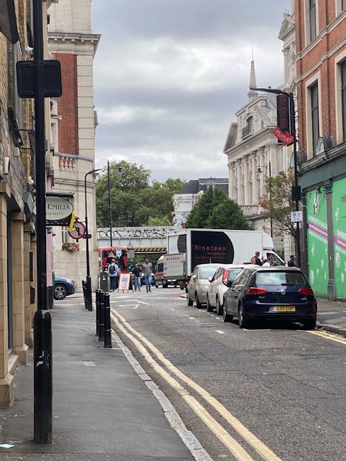 A narrow street in Maida Vale with parked cars on the right side and a black van next to them, part of a home relocation process. The van is stationary, and the street is lined with traditional brick buildings, some with shop signs, including one for Emilia Boutique. In the background, a white moving truck with the branding 'dinatee' is parked, with people beside it involved in loading or unloading furniture, boxes, and packing materials. The street features black bollards along the pavement, double yellow lines indicating parking restrictions, and a pedestrian crossing warning sign visible on the left side. Overhead, there is a cloudy sky, and a building with classical architecture and a spire can be seen further down the street. The scene captures the logistics of furniture transport and packing during a house move, with visible blankets and wrapping on some objects, under natural daylight typical of a daytime move managed by Maida Vale Man and Van.