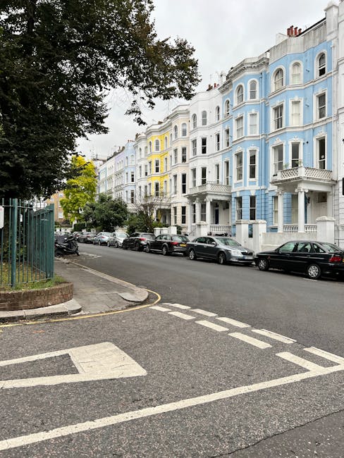 The image shows a street in Maida Vale with a row of four-storey Victorian-style residential buildings painted in pastel shades of blue, white, and yellow. The buildings feature bay windows, decorative cornices, and small front balconies. Parked along the curb are several cars, including sedans and hatchbacks, positioned on both sides of the narrow street. The street is lined with mature trees, providing some greenery and shade, with one large tree visible in the foreground on the left side of the image, partially obscuring the buildings. The pavement and street markings, including a pedestrian crossing with white dashed lines, are clearly visible in the foreground. The overall scene is during daytime with overcast weather, indicating typical conditions for home relocation or furniture transport planning in this area. This setting aligns with the professional removals services provided by Maida Vale Man and Van, who are experienced in managing tight street access and moving logistics in residential areas like Warwick Avenue Maida Vale. 