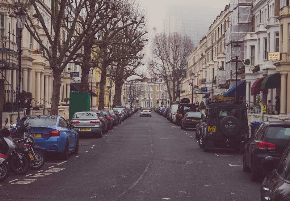 The image shows a street in Maida Vale with a row of four-storey Victorian-style residential buildings painted in pastel shades of blue, white, and yellow. The buildings feature bay windows, decorative cornices, and small front balconies. Parked along the curb are several cars, including sedans and hatchbacks, positioned on both sides of the narrow street. The street is lined with mature trees, providing some greenery and shade, with one large tree visible in the foreground on the left side of the image, partially obscuring the buildings. The pavement and street markings, including a pedestrian crossing with white dashed lines, are clearly visible in the foreground. The overall scene is during daytime with overcast weather, indicating typical conditions for home relocation or furniture transport planning in this area. This setting aligns with the professional removals services provided by Maida Vale Man and Van, who are experienced in managing tight street access and moving logistics in residential areas like Warwick Avenue Maida Vale. 
