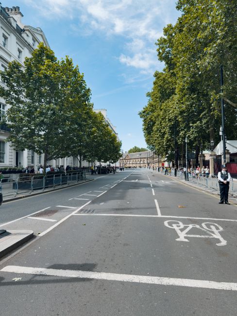 A quiet street in Maida Vale with a wide, paved road and designated bicycle lane marked with white paint. Flanking the street are mature green trees providing partial shade, and white residential buildings with ornate facades and balconies line the left side of the street. On the right side, a row of pedestrians is visible, some standing near the sidewalk, and there are streetlamps and signage along the pavement. The sky overhead is partly cloudy with patches of blue. In the foreground, a Maida Vale Man and Van vehicle is partially visible, parked next to the curb, with some furniture and packing boxes being loaded or unloaded during a home relocation or moving process, supported by the company's professional removals services, in accordance with Westminster Council moving rules and loading permits.